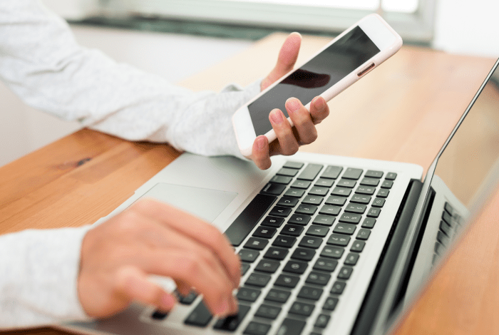 A person holding a smart phone and typing on a laptop with the other hand.