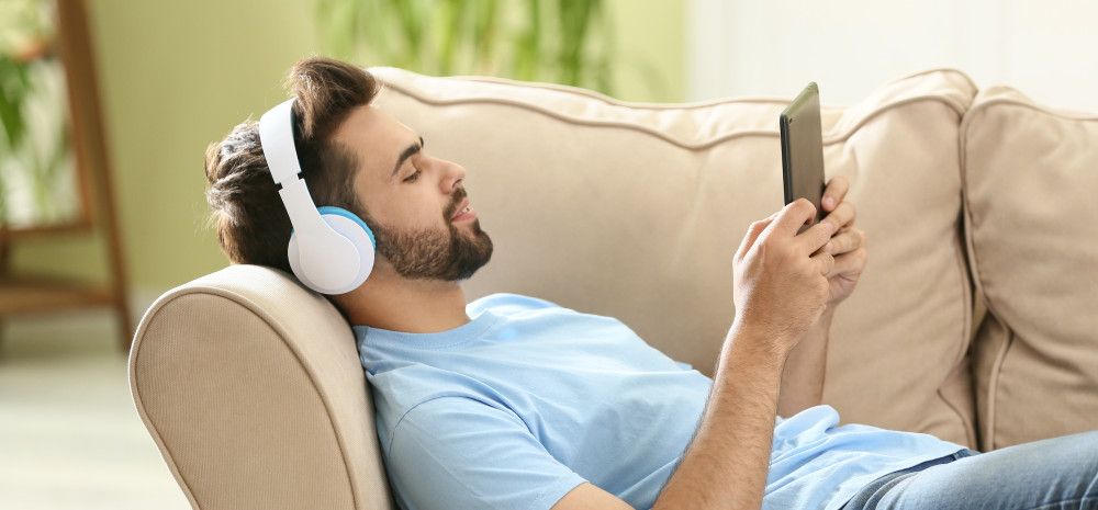 Man lying on a couch listening on headphones holding a mobile device.