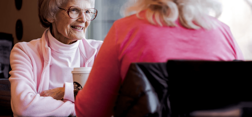 Two older women sitting at a table and chatting over a cup of coffee.