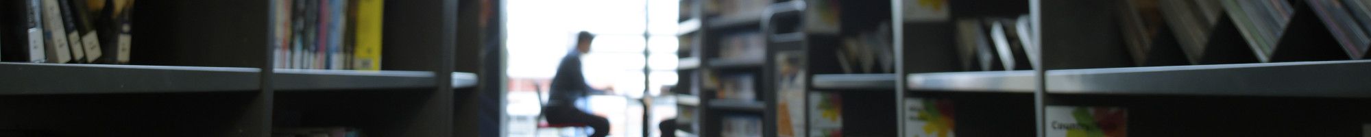 Image: Man sitting reading in a library, with bookshelves in the foreground.