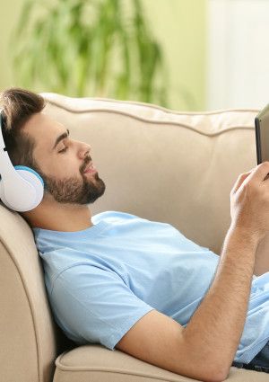 Man lying on a couch listening on headphones holding a mobile device.